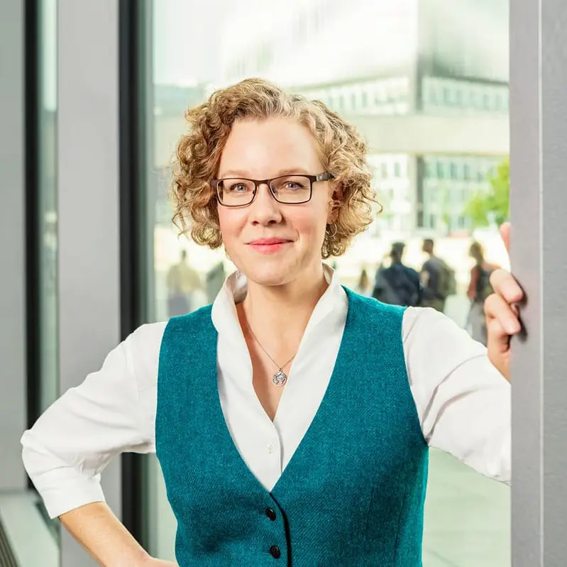 Julia Verlinden steht in einem modernen Gebäude. Im Hintergrund sind durch die Glasfront Menschen im Außenbereich zu sehen.