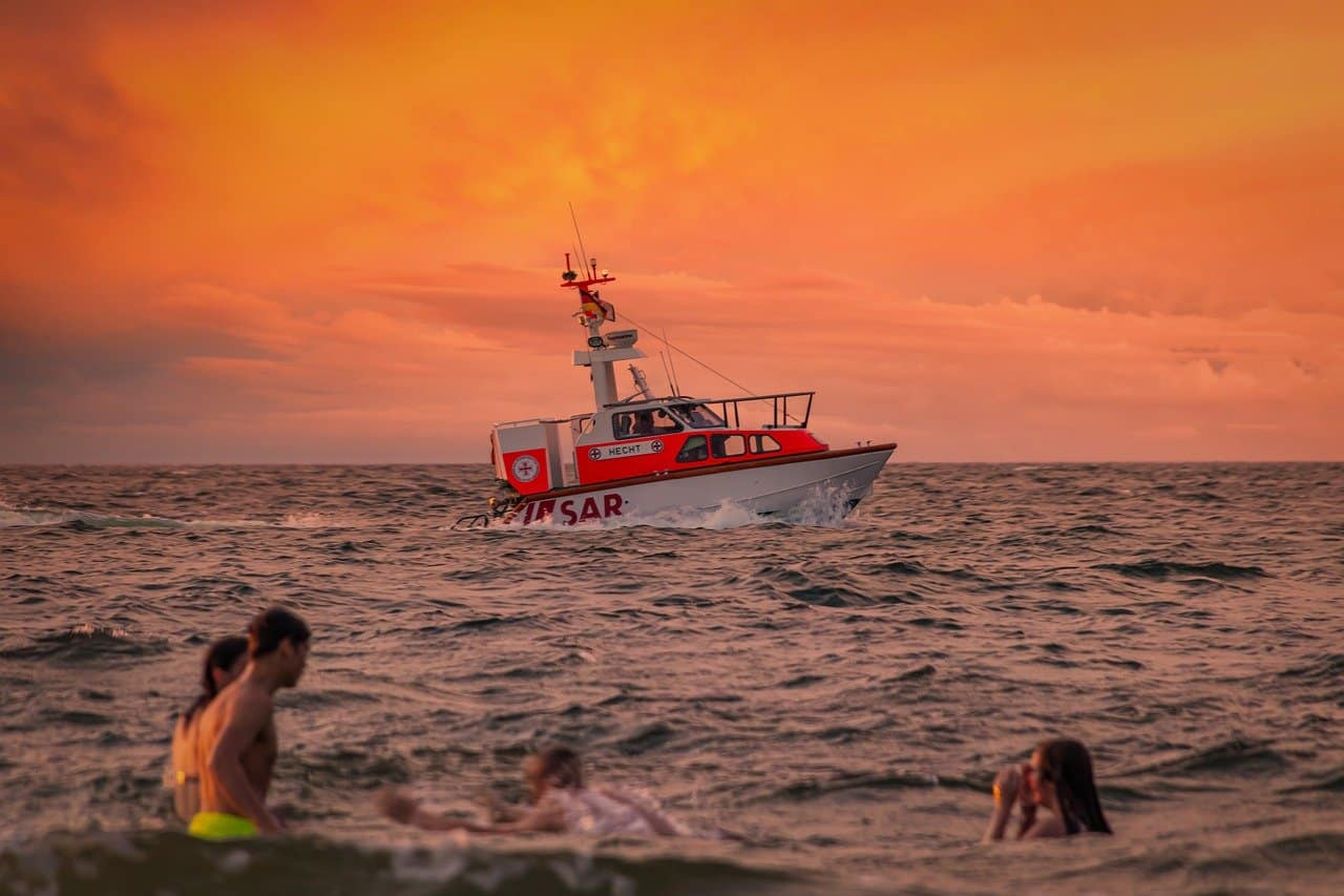 Badende und dahinter ein Rettungsboot auf dem Meer vor rotem Himmel